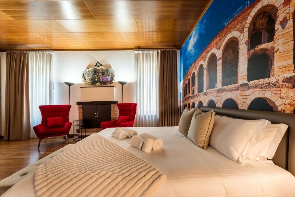 Ein Hotelzimmer mit einem großen Bett, zwei roten Sesseln, einem Kamin und einem Wandbild eines historischen Gebäudes.