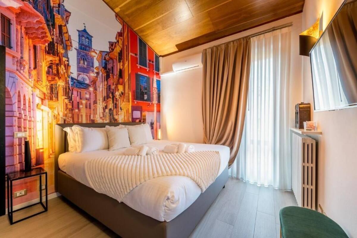 A hotel room with a large bed, a wall-mounted air conditioner, a bedside table, and a view of a cityscape through sheer curtains.