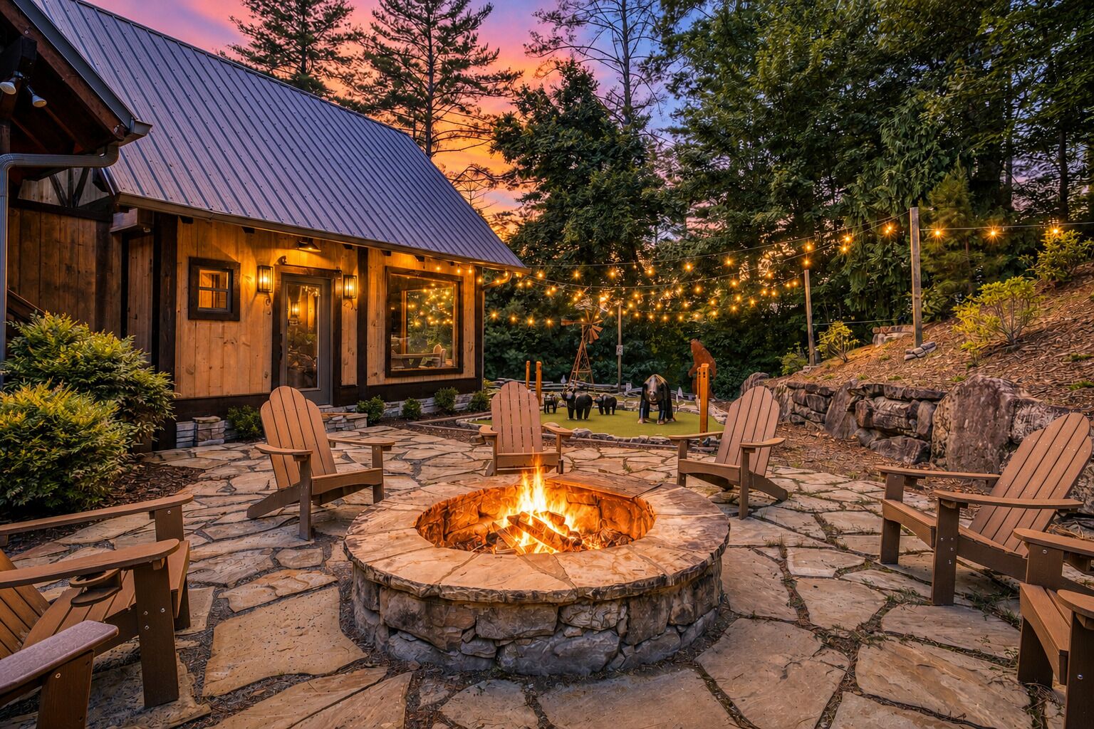 A stone fire pit with chairs, a wooden building with a metal roof, and string lights in a landscaped outdoor area.