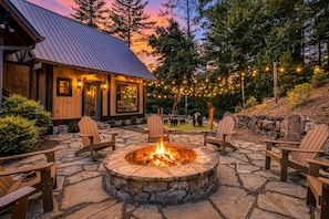 A stone fire pit with chairs, a wooden building with a metal roof, and string lights in a landscaped outdoor area. - Luxury Mountaintop Lodge: Mini Golf, Hot Tub, Views, Clubhouse, Pet Friendly (Gilmer County)