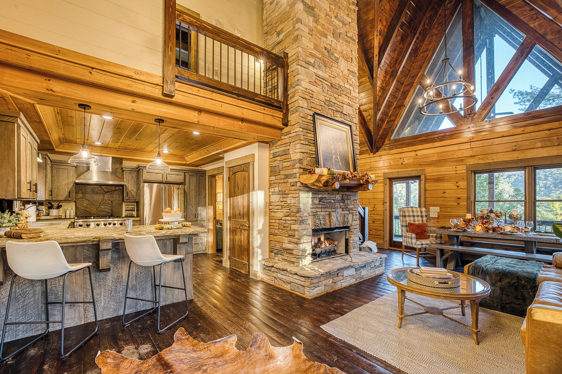 A spacious living area with a stone fireplace, wooden beams, and a large window overlooking a forest.