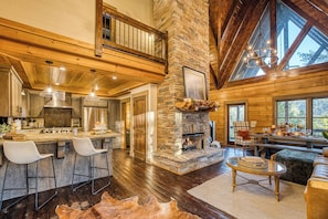 A spacious living area with a stone fireplace, wooden beams, and a large window overlooking a forest. - Luxury Mountaintop Lodge: Mini Golf, Hot Tub, Views, Clubhouse, Pet Friendly (Gilmer County)