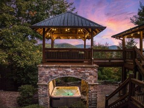 A hot tub under a wooden gazebo with a stone wall, surrounded by trees and hills. - Luxury Lodge: Mini Golf, Hot Tub, Views, Game Room, Pet Friendly (Gilmer County)