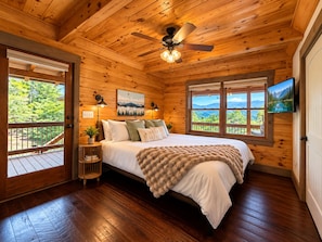 A wooden cabin bedroom with a bed, a ceiling fan, a window with a view, and a television on the wall. - Luxury Mountaintop Lodge: Mini Golf, Hot Tub, Views, Clubhouse, Pet Friendly (Gilmer County)