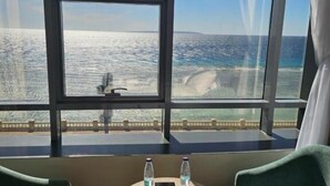 A room with a large window overlooking the sea, two green chairs, a small round table with a bottle and glass, and a bed.