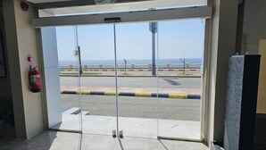 A glass door entrance with a fire extinguisher on the wall, a fire alarm, and a view of a road with a guardrail and palm trees.