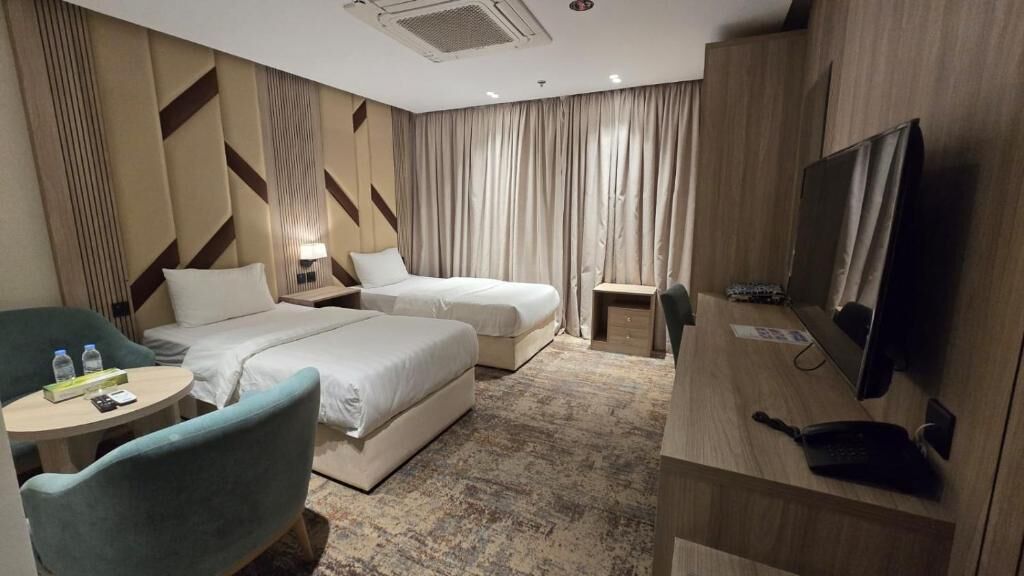 A hotel room with two beds, a TV, a desk, a chair, and a small table with water bottles.