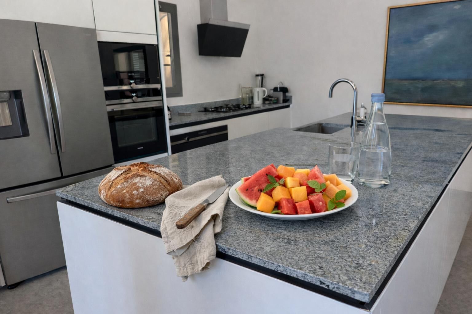 A modern kitchen with a granite countertop, a loaf of bread, a plate of fruit, and a water bottle.