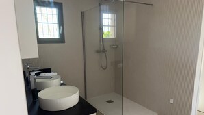Een moderne badkamer met een douchecabine van glas, een rond wastafeltje en een raam dat voor daglicht zorgt.