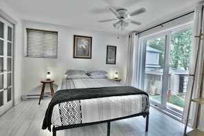 A bedroom with a bed, two bedside tables with lamps, a ceiling fan, and a large window with a view of the outdoors.