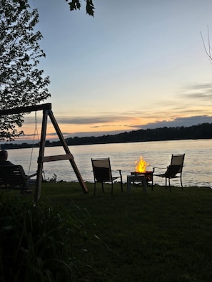 A lakeside setting with a swing, two chairs, and a small table with a fire.