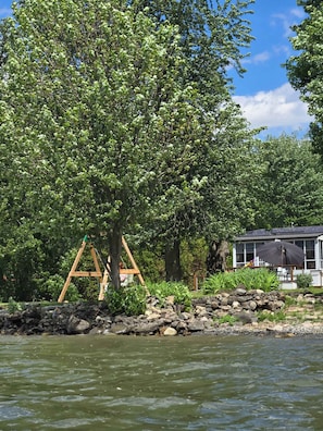 A lakeside view with a wooden swing set, a house with a covered patio, and a stone wall.