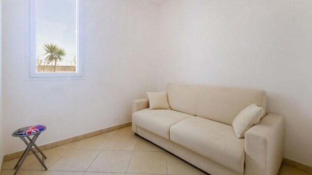 A room with a beige sofa, a small table with a flag, a window with a view of palm trees, and a ceiling light fixture. - Spacious and comfortable 90 sqm villa, ideal for up to 4 guests, equipped with all the comforts for a relaxing holiday in the heart of Salento, including private parking, a large garden, a covered patio, and a furnished terrace, perfect for enjoying rela (Torre dell'Orso)