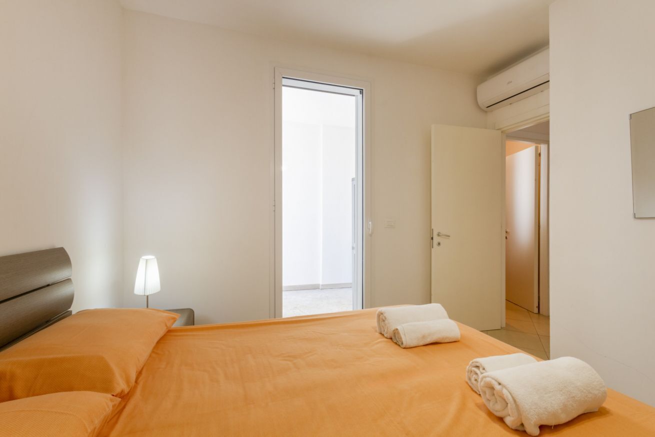 Hotellihuone, jossa on sänky, yöpöytä, lamppu, ovi ja ilmastointilaite.