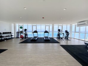 Un gimnasio con cintas de correr, elípticas y estantes de pesas, ventanas amplias y vista al mar.