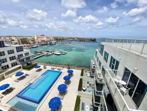 Una piscina en la azotea con sillones y sombrillas, un puerto deportivo con barcos y vistas a la ciudad.