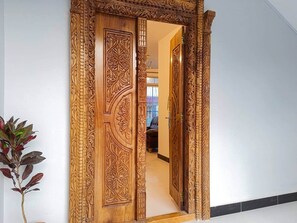Une porte en bois ornée de sculptures complexes, qui donne sur un couloir au sol de couleur claire et où se trouve une plante sur le côté gauche.
