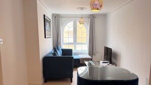 Een moderne woonkamer met een eettafel met glazen blad, een donkerblauwe bank en een groot raam met gordijnen.