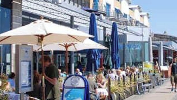 Een promenade aan zee met terrasjes, een man bij een kiosk en een helderblauwe lucht.