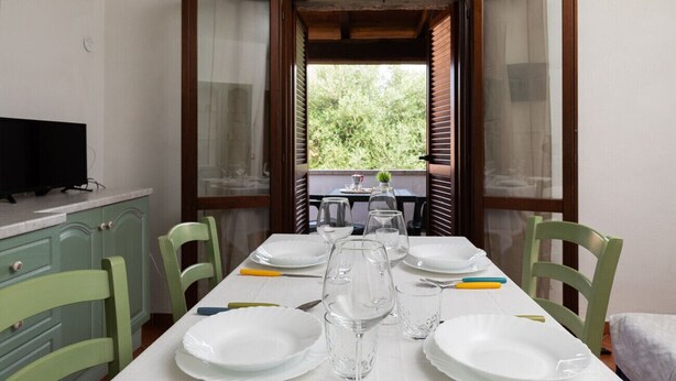 A dining area with a table set for four, chairs, a television, and a view of greenery outside.