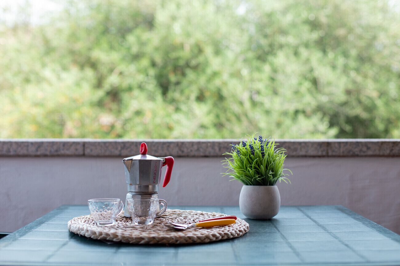 Een tafel met een koffiezetapparaat, twee glazen, een lepel en een potplant op een geweven onderzetter.
