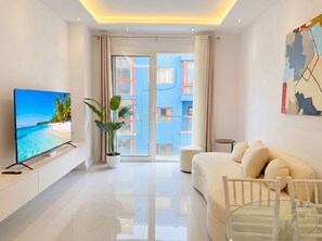 Ein modernes Wohnzimmer mit Flachbildfernseher, Topfpflanze, Sofa, Glastisch und Blick auf ein blaues Geb äude durch die Glastür.