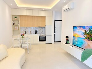 Ein modernes Wohnzimmer mit einer Couch, einem Fernseher, der eine Strandansicht zeigt, einer Küche mit einer Kaffeemaschine und einer Tür zu einem weiteren Raum.