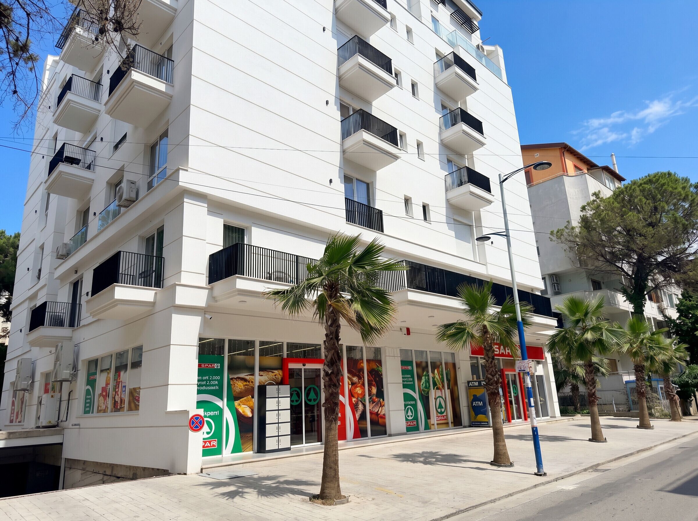 A multi-story building with balconies, a storefront with advertisements, and palm trees.