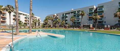 A swimming pool with clear blue water, surrounded by palm trees and modern apartment buildings.