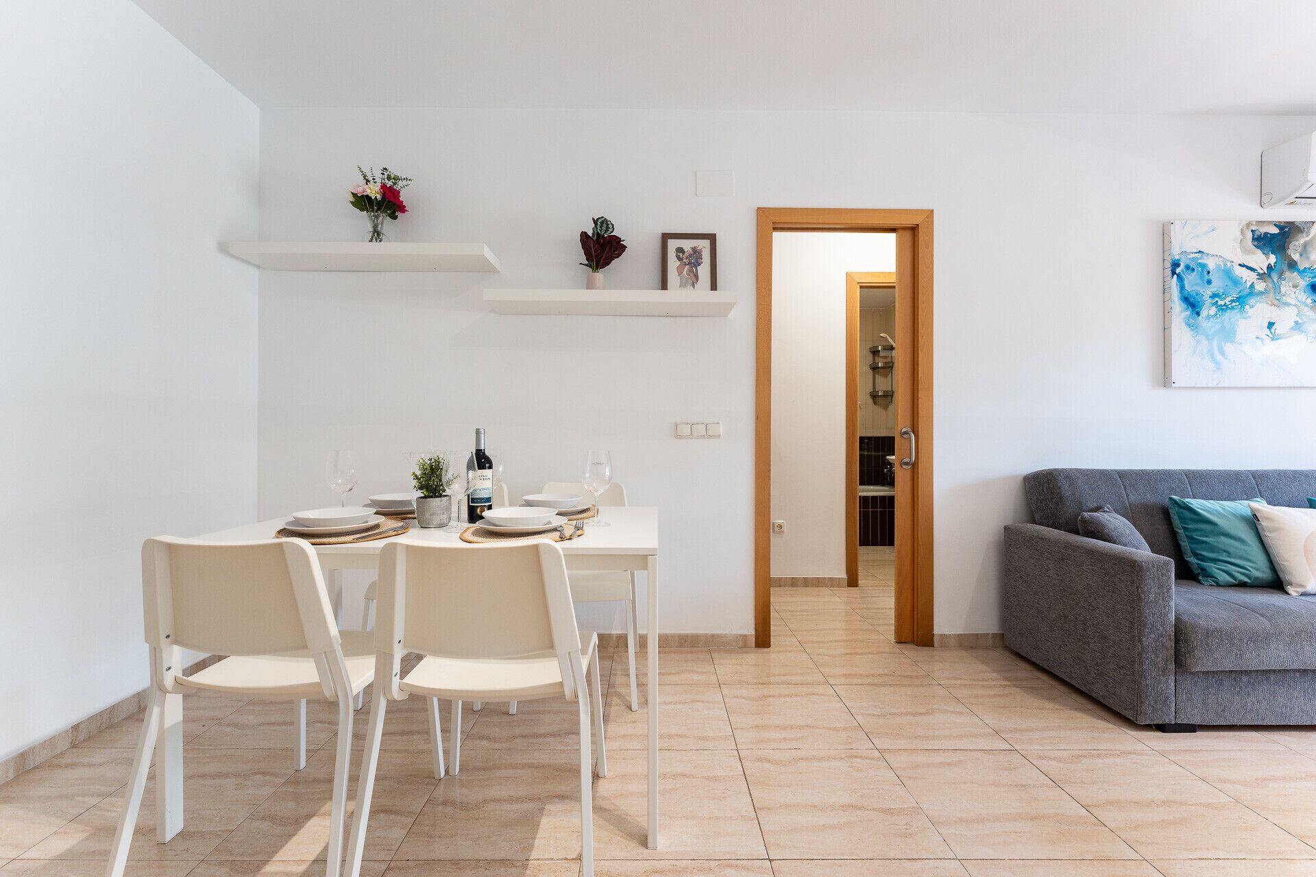 A dining area with a table set for four, a grey sofa, and a kitchen visible through an open door.