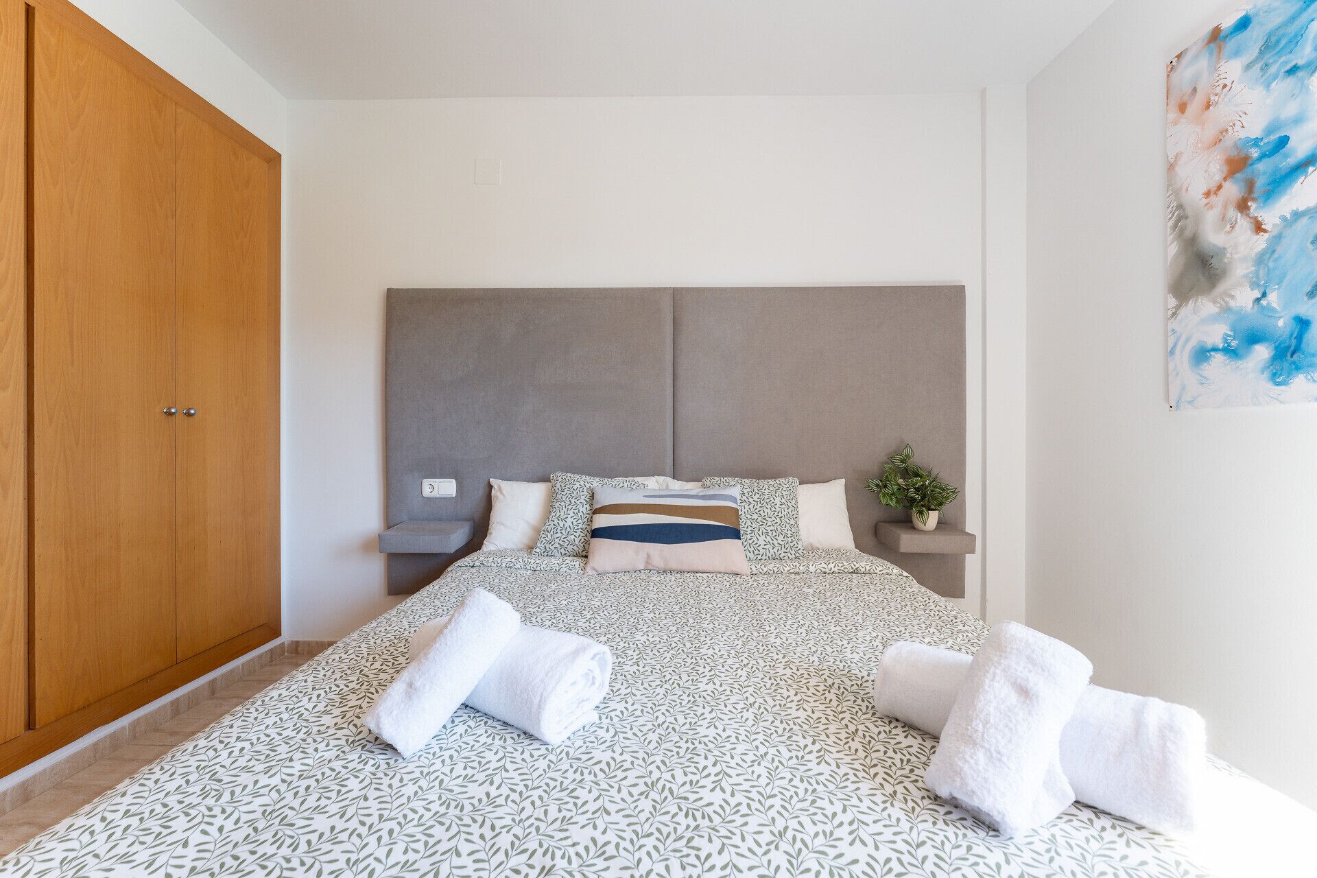 A bedroom with a large wardrobe, a bed with patterned bedding, a headboard, bedside tables, and a wall-mounted plant.