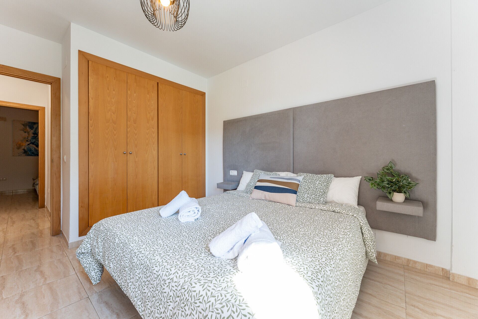 A bedroom with a bed, a wooden wardrobe, a grey headboard, and a small plant.