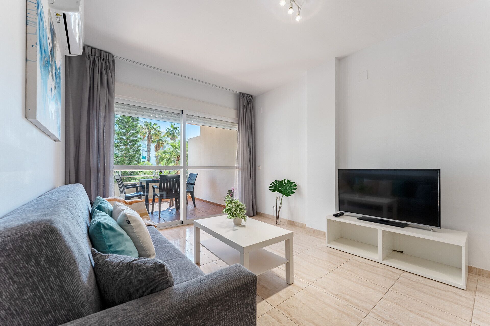 Un salon moderne avec un canapé, une table basse, un téléviseur posé sur un meuble et un balcon offrant une vue sur des palmiers.