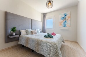 Une chambre moderne avec un grand lit, une plante sur la table de chevet, une étagère fixée au mur, un luminaire au plafond et une fenêtre avec des rideaux.