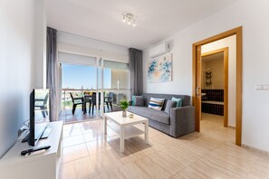 A modern living room with a sofa, coffee table, and TV. There is a balcony with a view, and an open door leading to a bathroom. - AlmeriaSuite Palm Beach Golf & Pool 6301 (El Ejido)