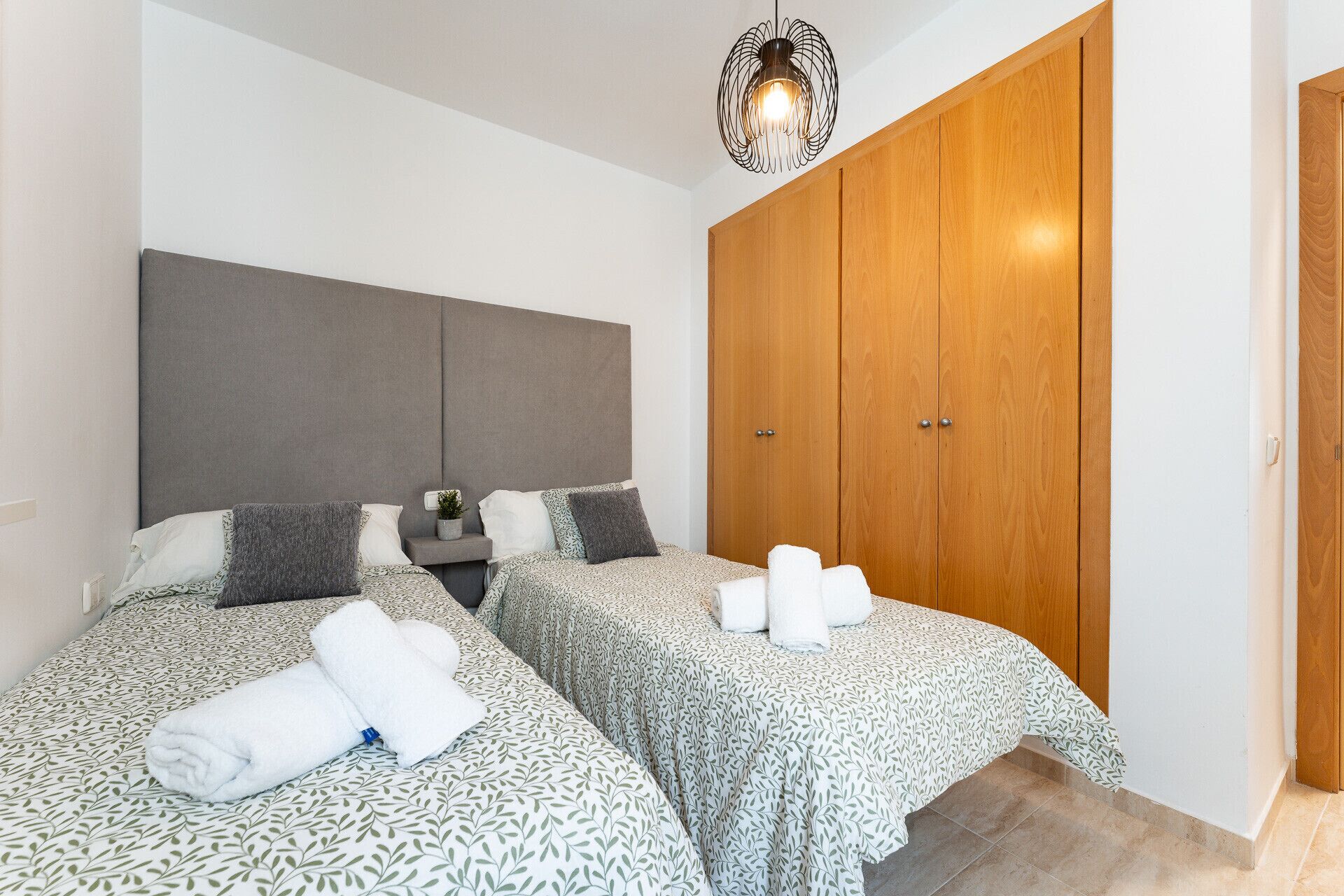 A bedroom with two beds, a grey headboard, a wooden wardrobe, and a hanging light fixture.