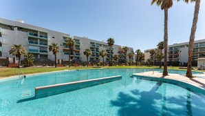 A swimming pool with clear blue water, surrounded by palm trees and a multi-story building with balconies.