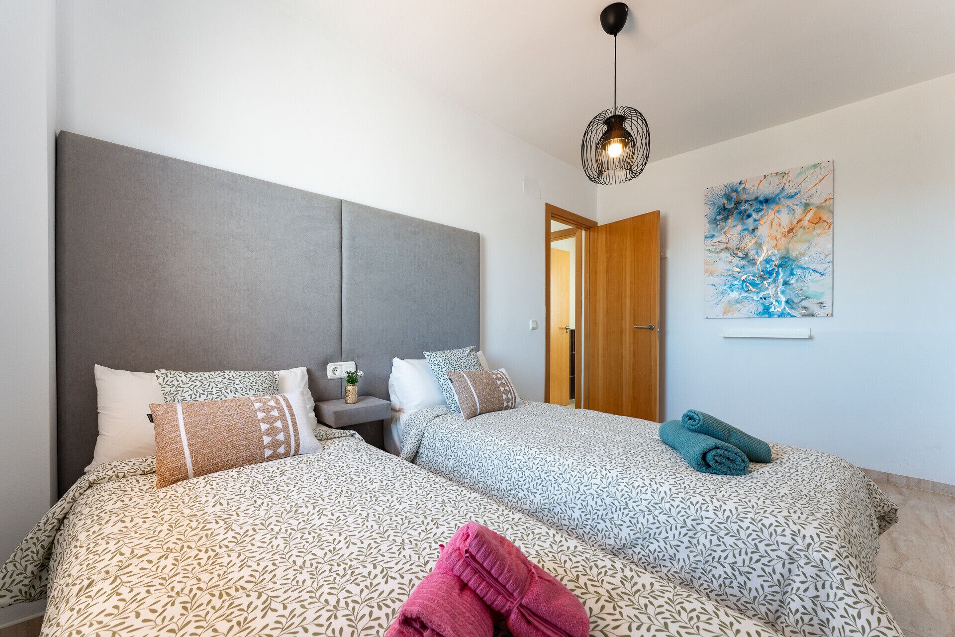 A double bed with patterned bedding, a bedside table with a plant, a modern pendant light, and a door leading to another room.