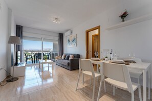 A modern living room with a sofa, dining table, and chairs, a balcony with a view, and a painting on the wall. - Almeria Suite 7305 Palm Beach (El Ejido)