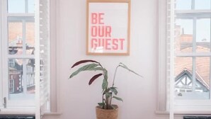 Quarto com piso de madeira, uma pequena mesa com uma planta, um quadro e uma janela com persianas.