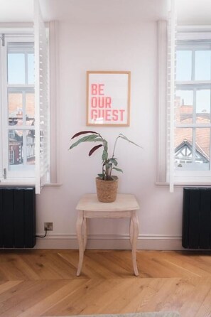 Ein Zimmer mit Holzboden, einem kleinen Tisch mit Pflanze, einem gerahmten Schild und einem Fenster mit Jalousien.
