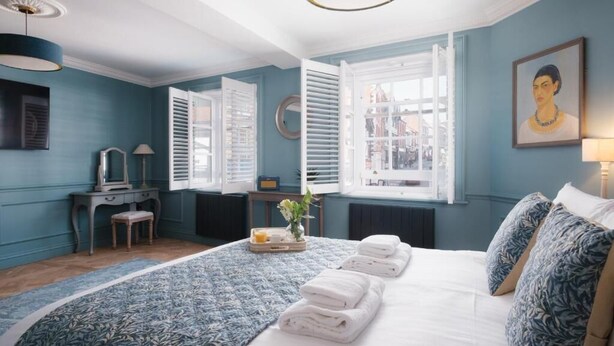 Um quarto com cama, escrivaninha, cadeira, janela com persianas e um quadro na parede.