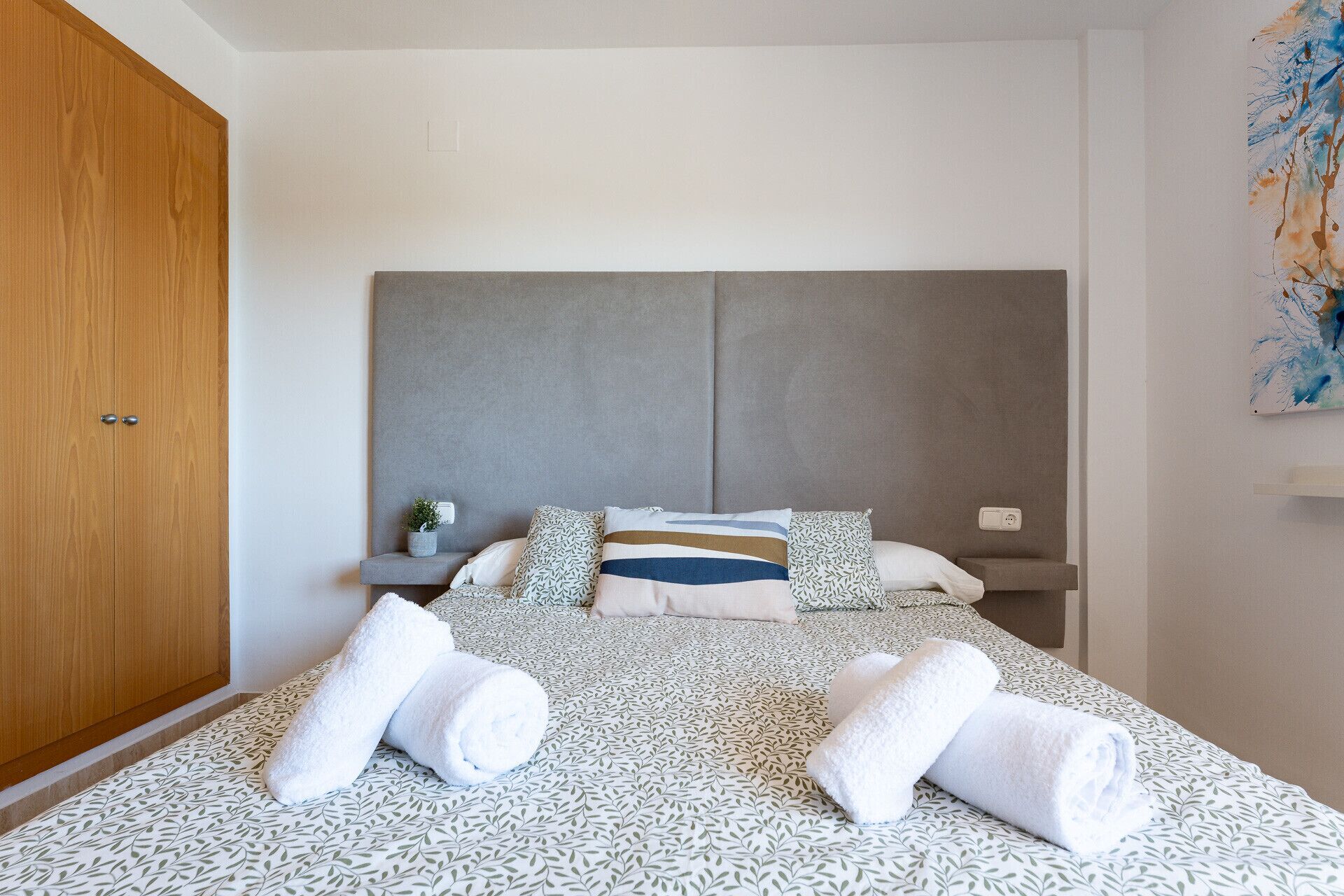 A bedroom with a bed, pillows, a nightstand, a potted plant, and a wooden wardrobe.