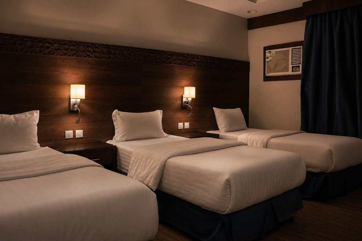 A hotel room with three beds, dark wood headboard, wall-mounted lights, and a window with curtains.