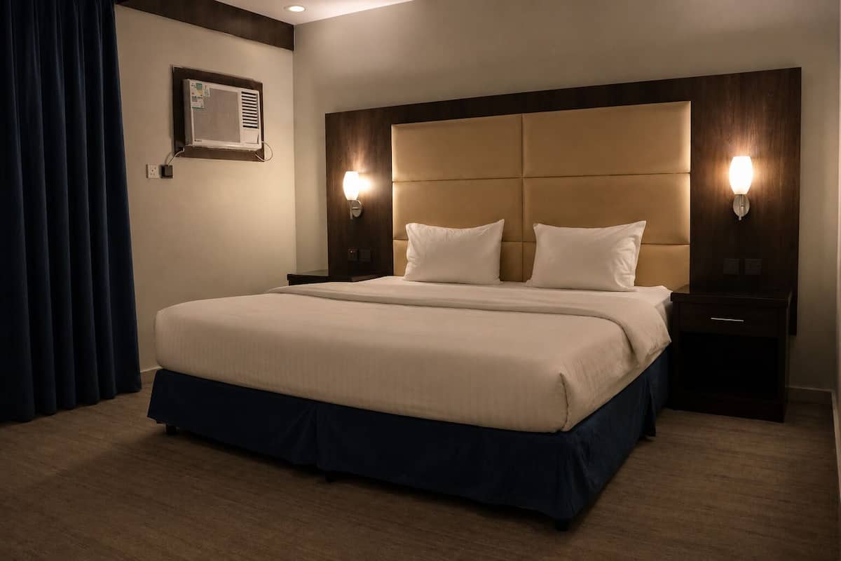 A hotel room with a large bed, two bedside tables, a wall-mounted air conditioning unit, and a dark curtain.