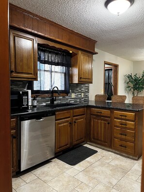 A kitchen with wooden cabinets, a stainless steel dishwasher, a tiled backsplash, and a black rug.