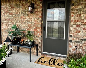 A brick wall, a door with a window, a black lantern, a small table with plants, and a 'hello' mat.