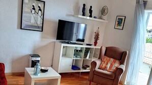 Een woonkamer met een televisie, witte planken, een rode fauteuil, een kleine tafel en een raam met gordijnen.