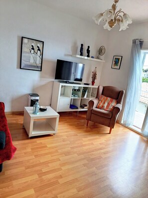 Ein Wohnzimmer mit Fernseher, weißen Regalen, einem roten Sessel, einem kleinen Tisch und einem Fenster mit Vorhängen.