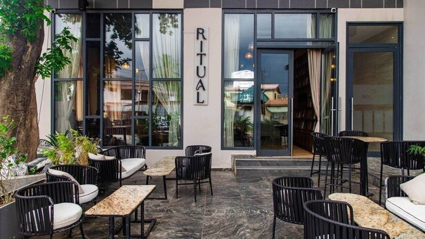 An outdoor seating area with tables and chairs, a tree, and a building with large windows and a sign that reads 'RITUAL'. - Solaris Premier 1BR | Osu • Pool • Gym (Accra)